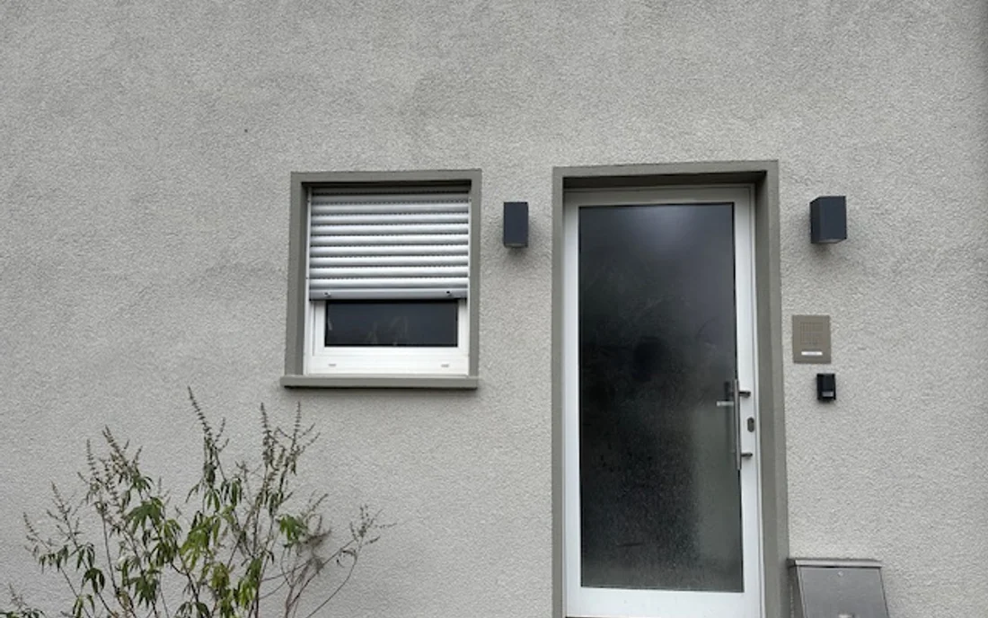 Hauseingang mit weißer Haustür, Fenster und Rollladen an einer grauen Putzfassade
